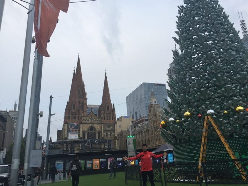 Dani en Melbourne chico mirando a cámara y detrás los edificios de Melbourne y el árbol de Navidad con el cielo nublado